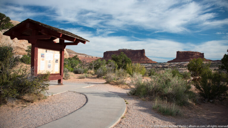 A paved path curves beside an information kiosk toward distant sandstone buttes.