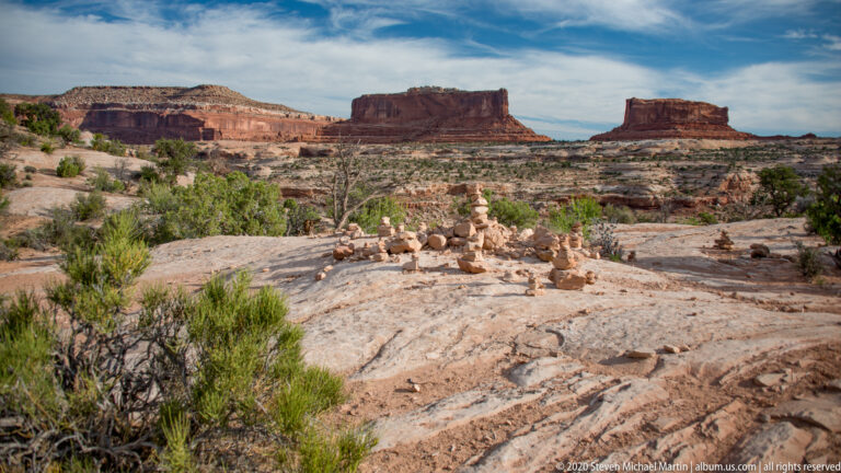 Scattered stone cairns and shrubs occupy a sandstone overlook below distant buttes.