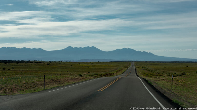 A straight highway crosses open grassland toward distant jagged mountains.
