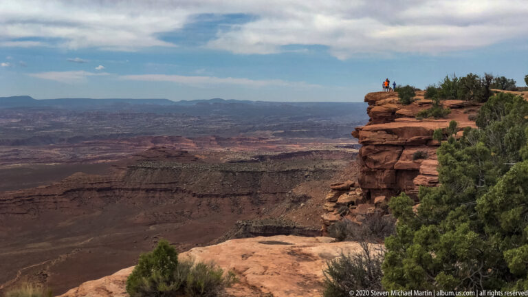 Two visitors stand on a rocky overlook above a deep canyon.