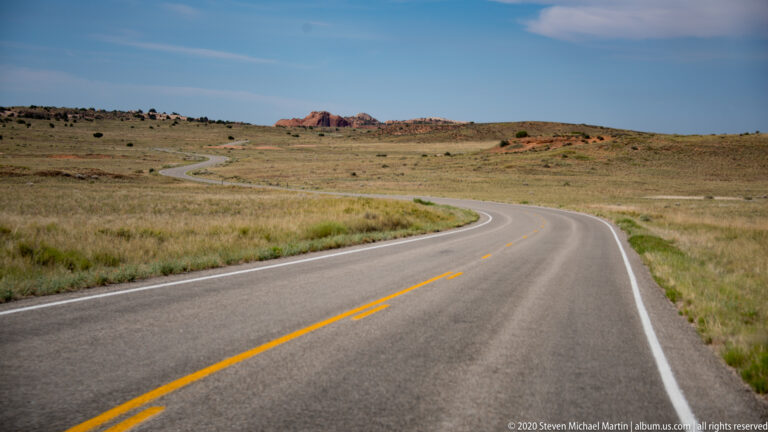 A paved road curves through grassland toward low red rock formations.