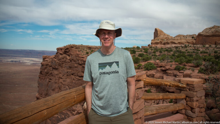 A man in a gray shirt and brimmed hat stands by a wooden overlook railing.