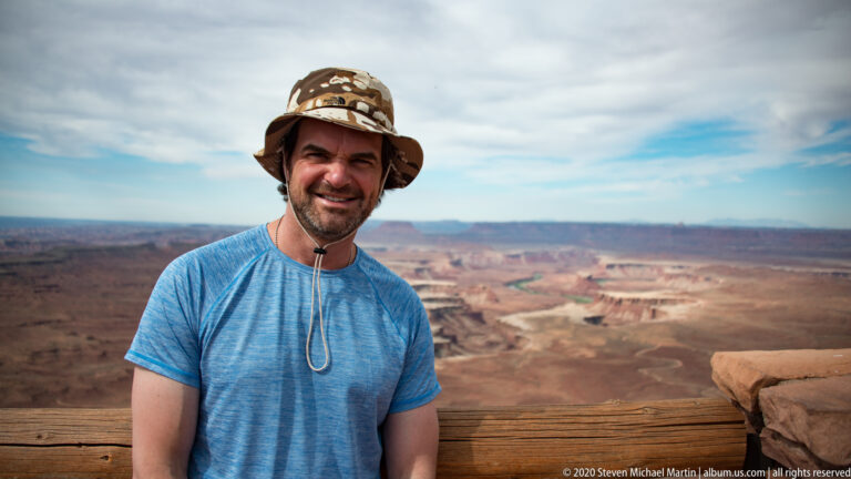 A man in a blue shirt and patterned hat stands at a canyon overlook.