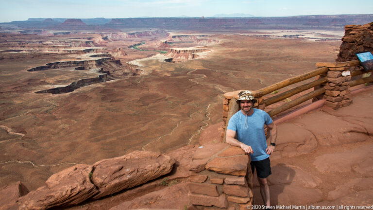 A visitor stands beside a timber overlook railing above a broad, layered canyon landscape.