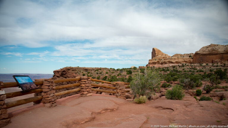 A timber-and-stone railing and interpretive panel edge a rocky overlook with junipers and a sandstone tower.