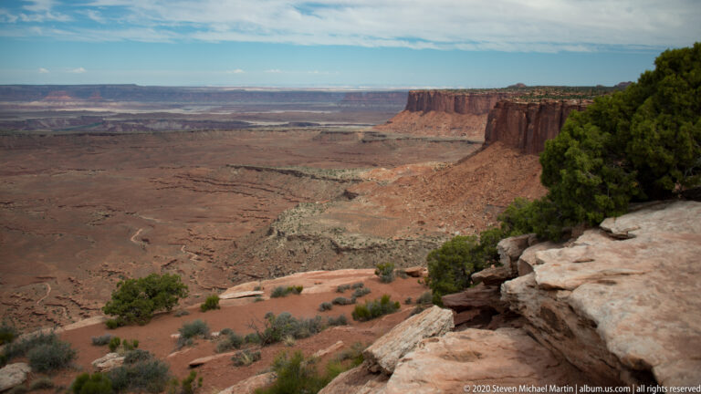 Pale sandstone slabs and juniper frame a long red escarpment above a distant canyon basin.