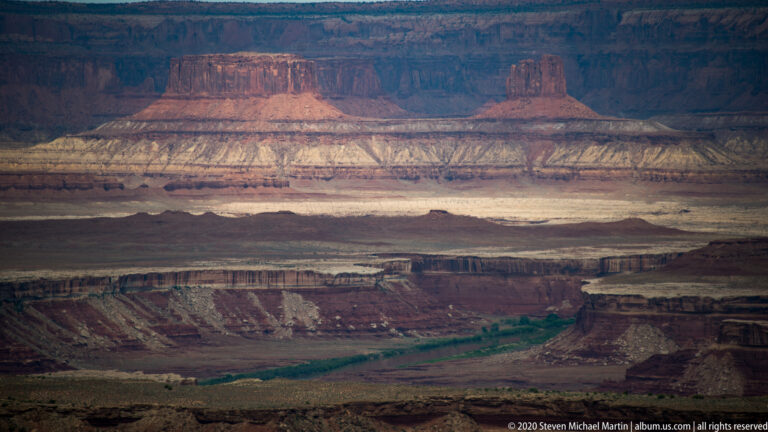 A banded red mesa rises beyond shadowed canyon shelves, with a pale river corridor far below.