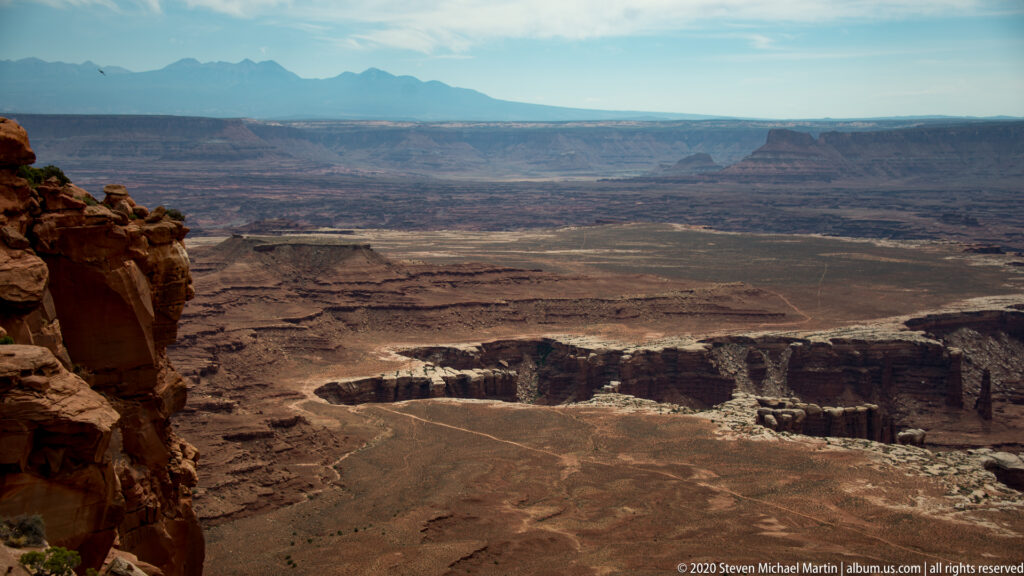 A shadowed cliff edge frames a vast view of mesas and branching canyons beneath distant blue mountains.