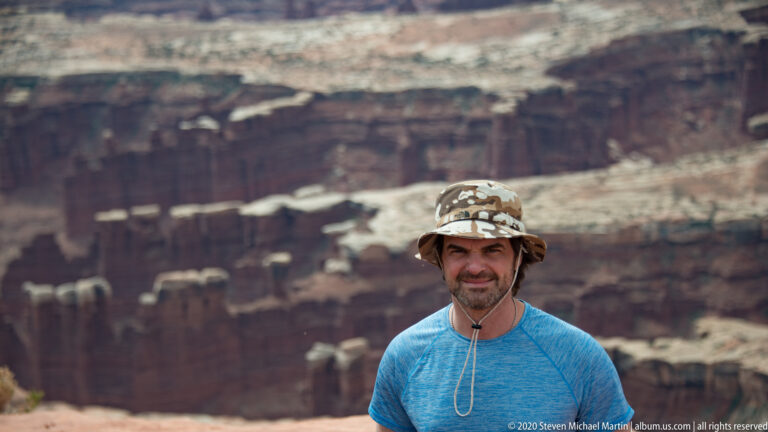 A visitor in a patterned hat and blue shirt poses before a softly blurred layered canyon.