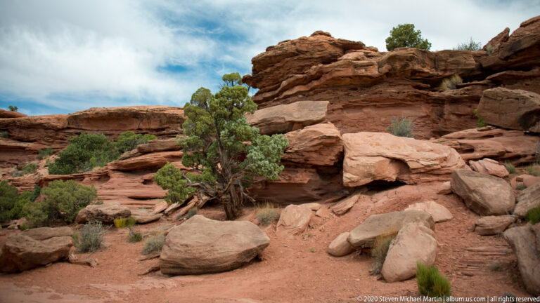 Layered red sandstone boulders rise among junipers and shrubs on a sandy slope.