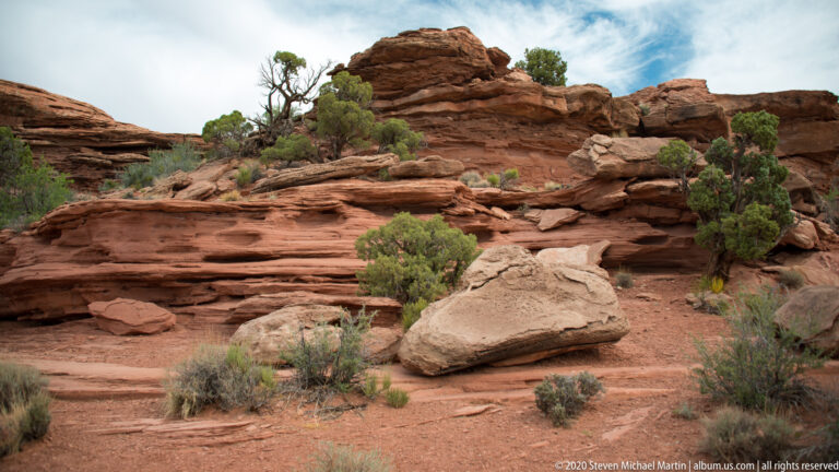 A projecting sandstone slab and rounded boulders sit among junipers and shrubs.