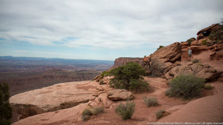 A lone visitor stands on a rocky rim trail above a broad layered canyon and escarpment.