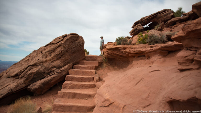 Rough stone steps climb between red sandstone slabs toward a visitor and open canyon view.