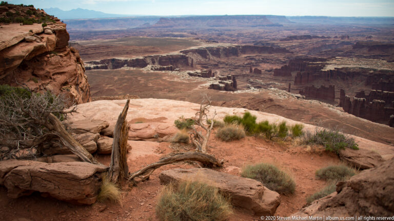 Pale rim boulders overlook branching canyons and flat-topped mesas stretching to a hazy horizon.