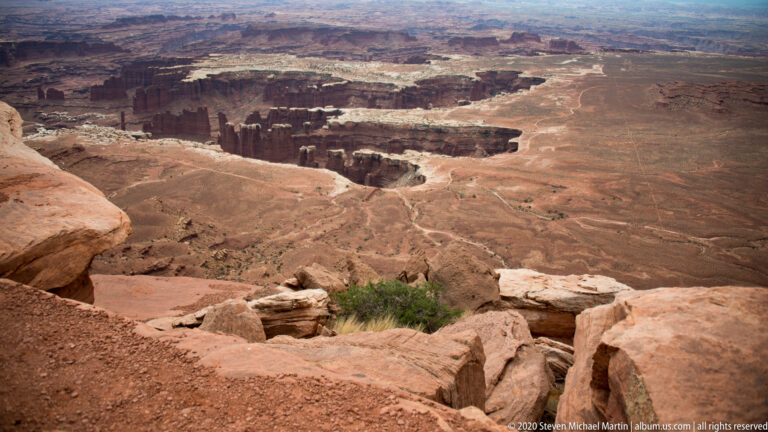 A large red mesa rises above pale rock bands and darker canyon shelves, with distant plateaus behind.