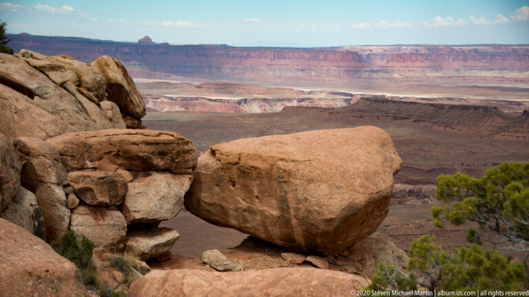 A flat-topped red butte and a narrow rock spire rise above a rubble-strewn canyon basin.