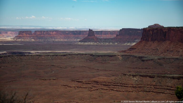 Red, cream, and brown rock bands cross a broad sparsely vegetated plateau beneath blue sky.