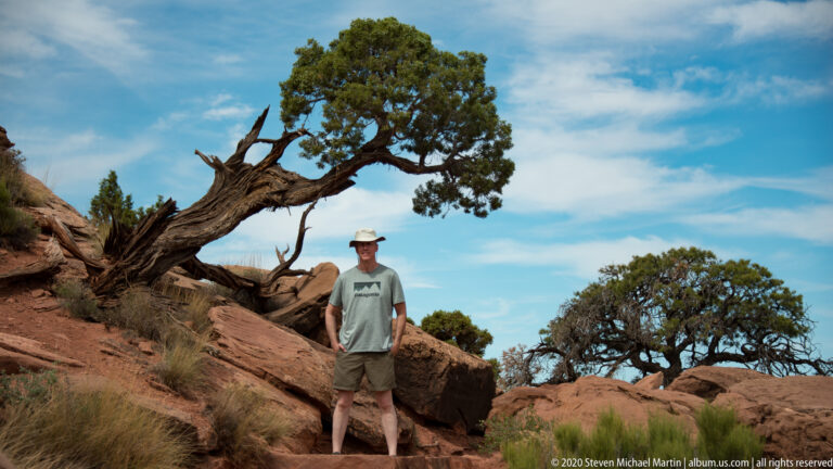 A small hiker stands among red boulders above a deep canyon, facing a sheer flat-topped mesa.
