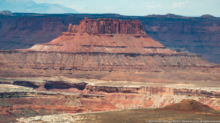 A large red mesa rises above pale rock bands and darker canyon shelves, with distant plateaus behind.