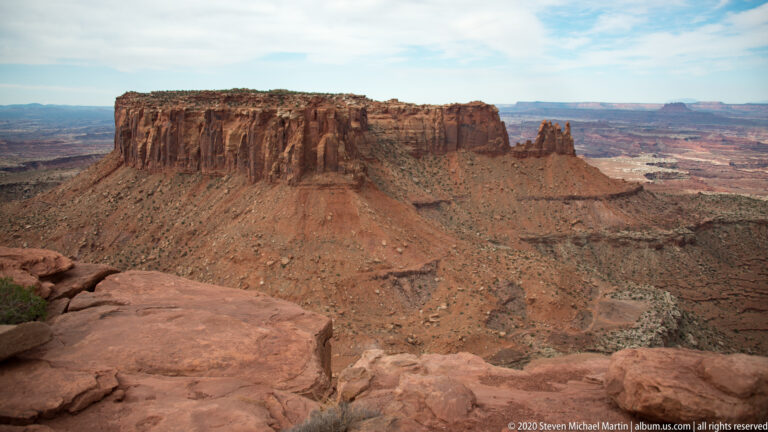 A flat-topped red butte and a narrow rock spire rise above a rubble-strewn canyon basin.