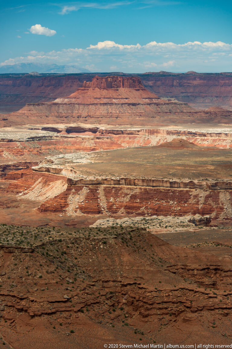 Red, cream, and brown rock bands cross a broad sparsely vegetated plateau beneath blue sky.