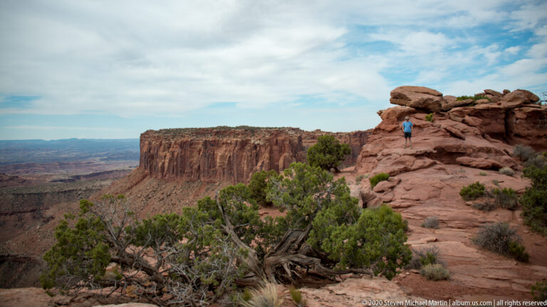 A small hiker stands among red boulders above a deep canyon, facing a sheer flat-topped mesa.