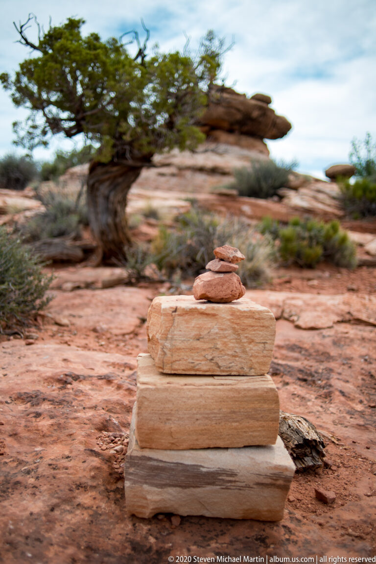 A small cairn of flat stones stands in focus before blurred red rocks and a juniper tree.