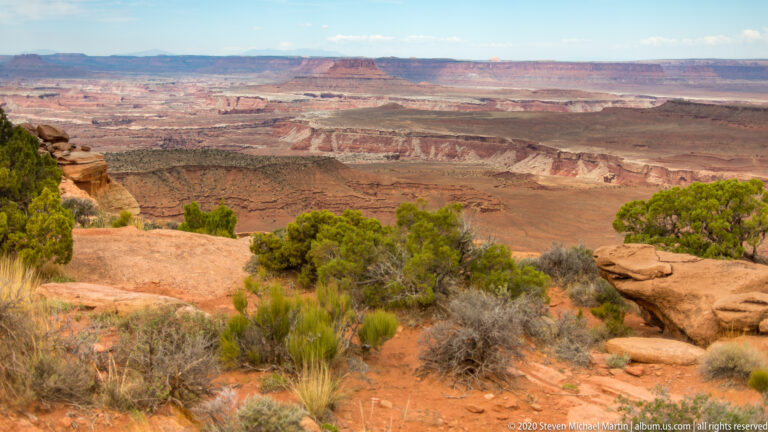 A red rock ledge and shrubs frame a deep basin of layered mesas and distant plateaus.