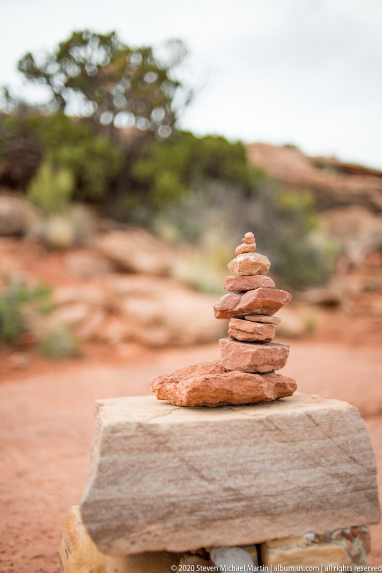 Flat reddish stones form a small cairn on a pale slab, with the desert background softly blurred.