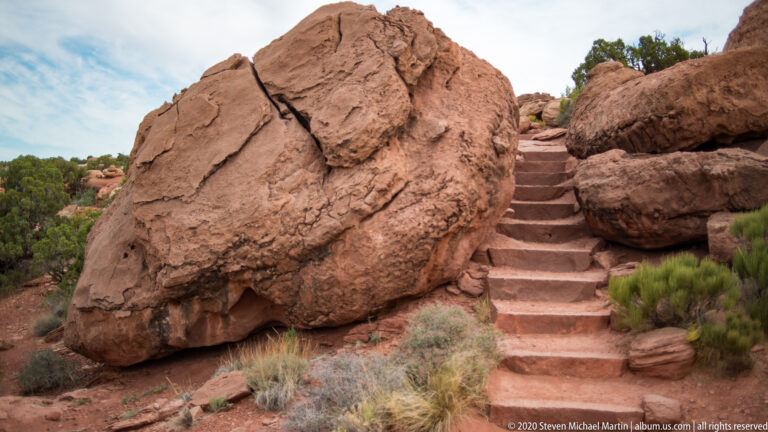 Sandstone steps climb through a narrow gap between large boulders and sparse grasses.