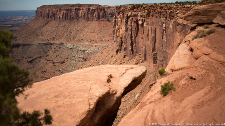 A small hiker stands on a pale ledge beneath a tall banded cliff and deep canyon walls.