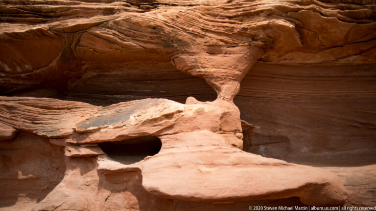A narrow sandstone pedestal supports a broad layered overhang above flat ledges in a shaded alcove.
