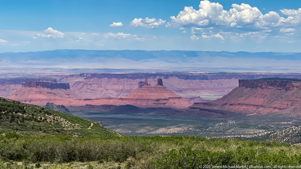 A central butte rises beyond a green foreground slope, with red mesas and blue ridges behind it.