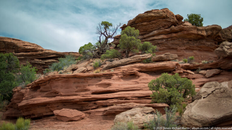Junipers grow among broken horizontal sandstone shelves beneath a taller rocky outcrop.