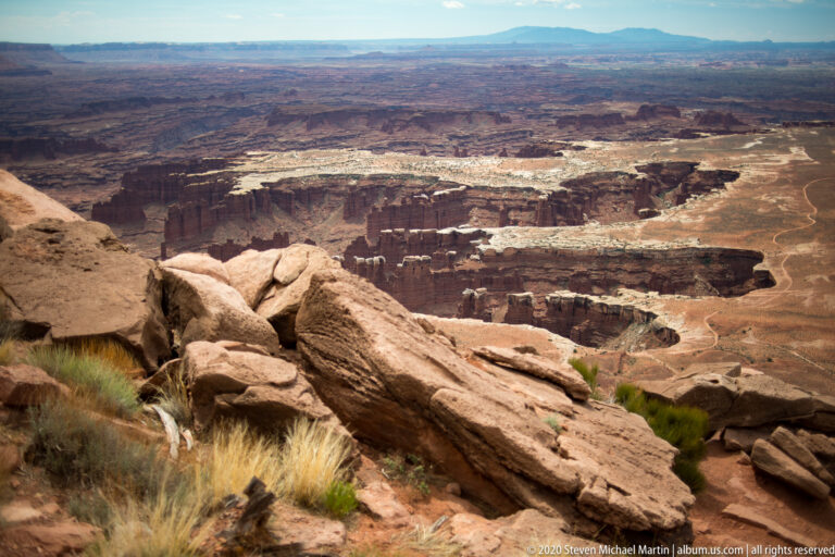 Rounded boulders and dry grasses overlook a deep canyon of cliffs, mesas, and ravines.