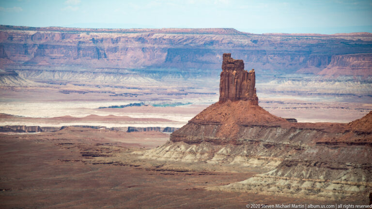 A solitary rock tower stands on a low pedestal amid pale basins and distant layered cliffs.