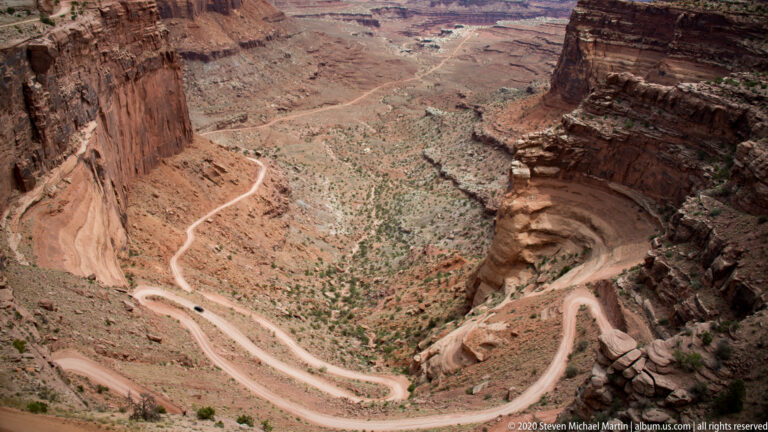 A pale dirt road zigzags down a steep canyon slope between high sandstone walls.