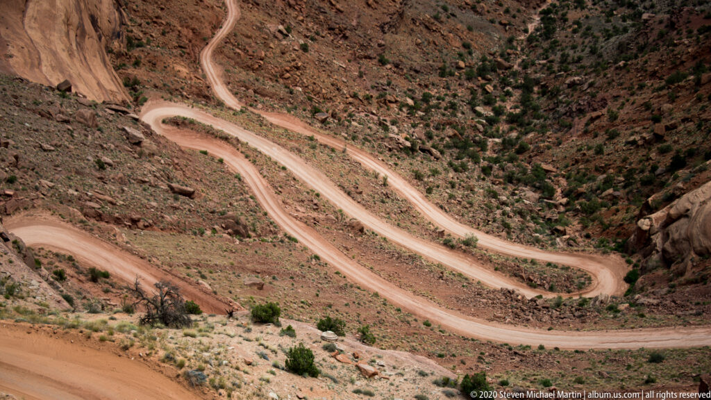 A pale dirt road forms a tight hairpin bend across a steep rocky slope dotted with scrub.