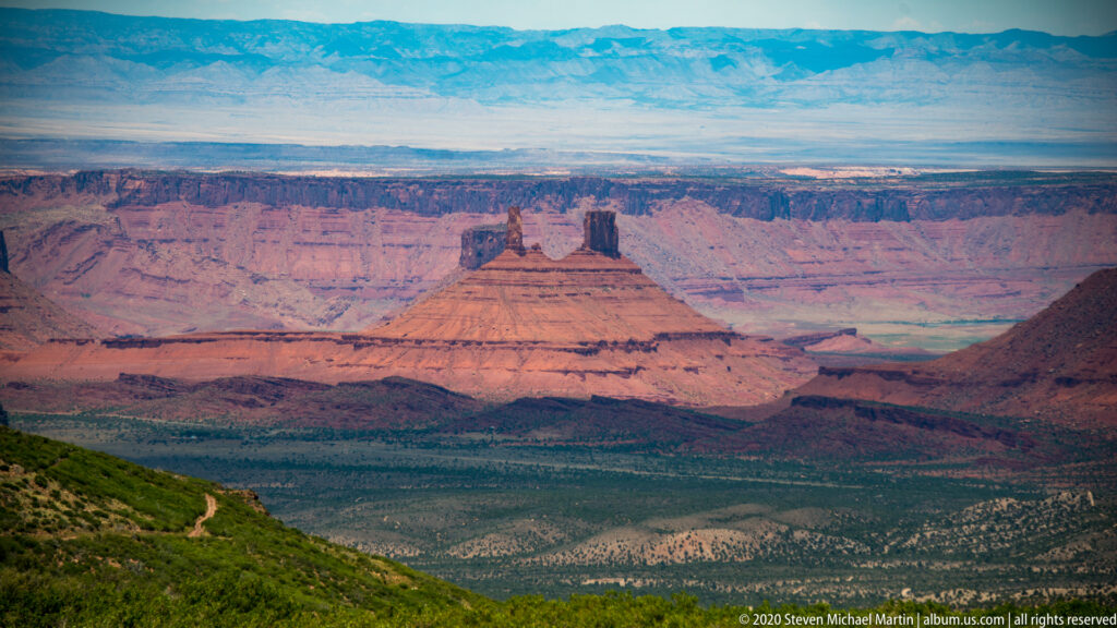 A flat-topped sandstone tower stands above a broad mesa and a greener, tree-covered valley.