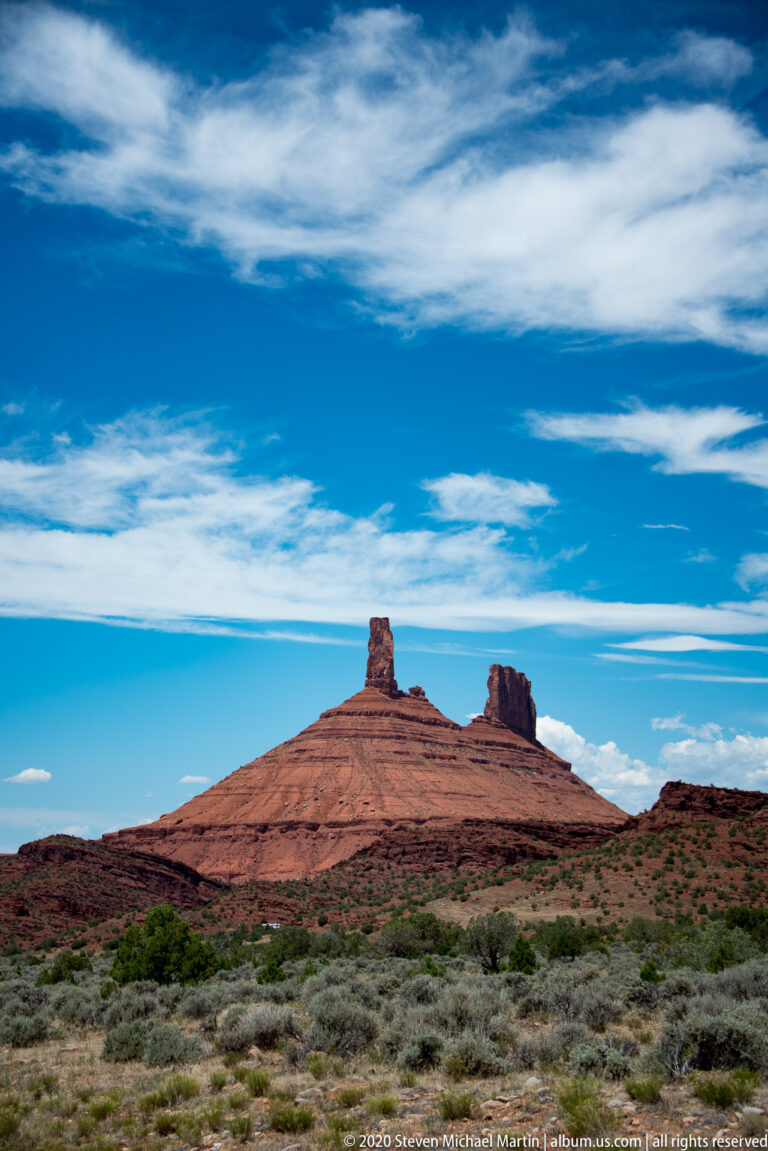 Two sandstone pinnacles break the line of a long ridge above foreground sagebrush beneath streaked clouds.