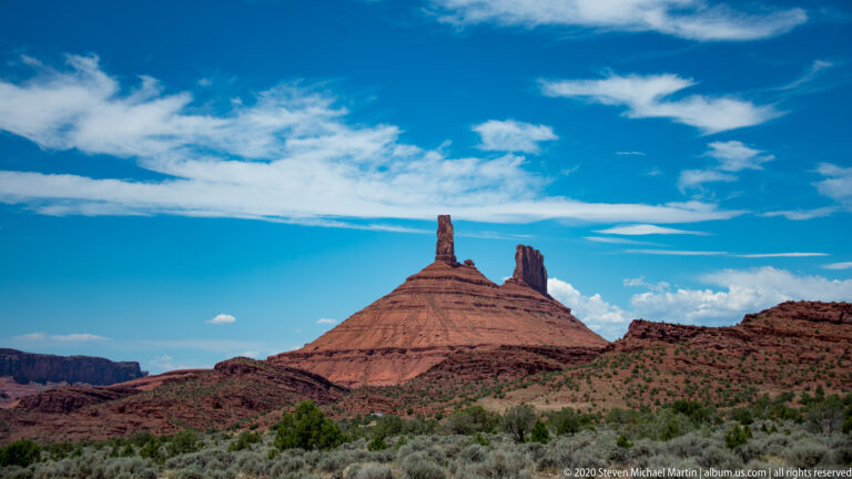A slender spire and a blockier sandstone tower rise above a sloping red rock base in the distance.