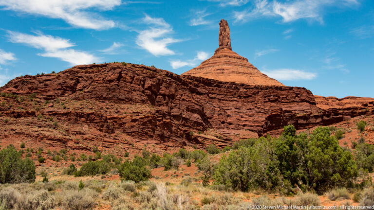 A narrow sandstone spire rises from a layered sloping mesa above sage and juniper.