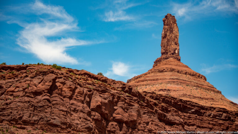 A tall sandstone spire stands on a stepped conical base partly obscured by a foreground ridge.