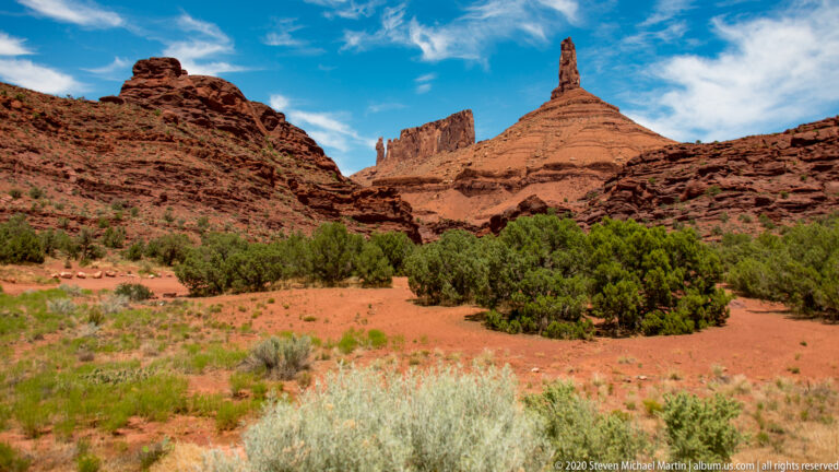 A needle-like spire and blocky sandstone tower overlook green trees in a valley beyond foreground sagebrush.