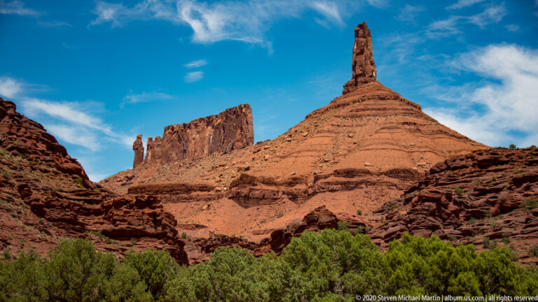 A narrow sandstone spire stands in front of a broad vertical tower above a strip of green vegetation.