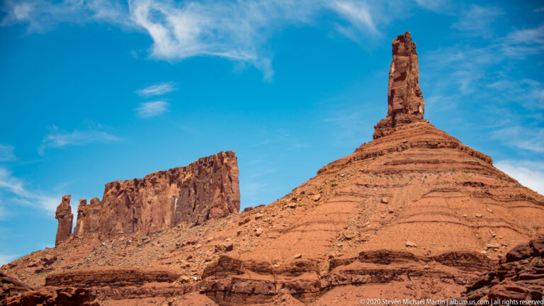 A broad sheer sandstone tower and a slender spire stand side by side against blue sky.