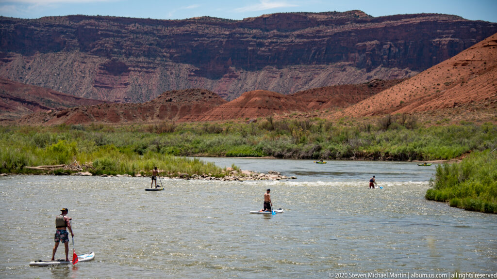 Three paddleboarders move across the Colorado River, with a scrubby bank and canyon wall behind them.