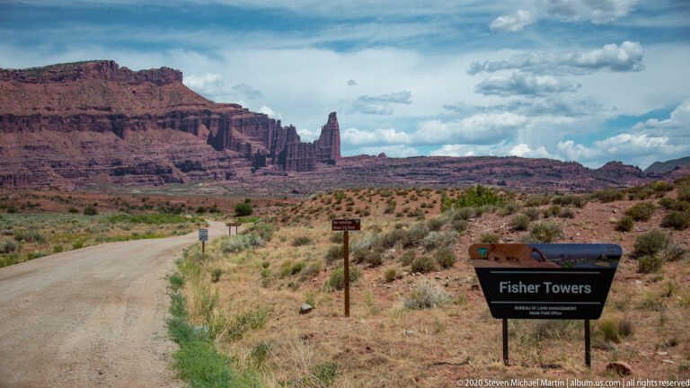 A gravel road approaches the red sandstone fins of Fisher Towers beside a roadside sign.