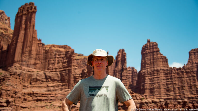 A man wearing a gray graphic T-shirt and brimmed hat stands before towering sandstone fins.