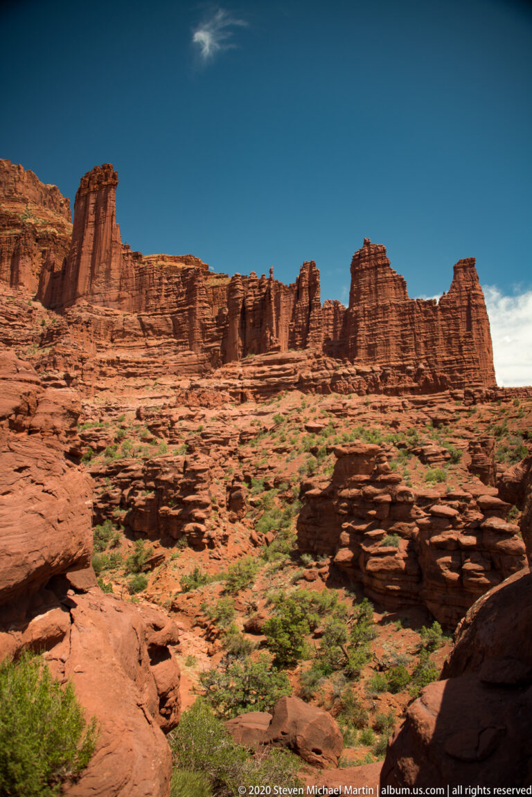 Sandstone fins and pinnacles rise beyond a sandy red foreground dotted with low shrubs.